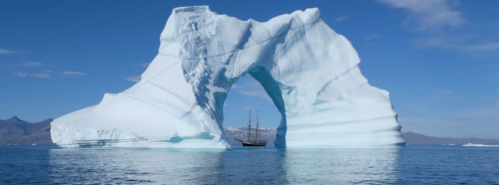 gigantischer Eisberg mit Loch in der Mitte - man sieht ein Segelschiff vorbeifahren